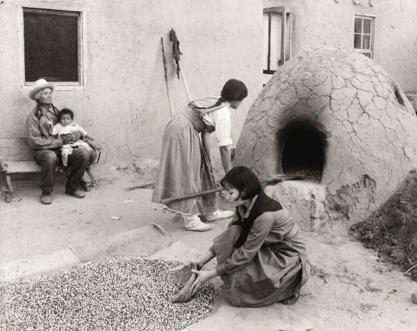 Historic black and white photo of two women tending to an outdoor indigenous oven with an elder holding a baby in the background