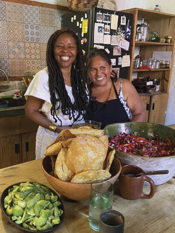 Two women smiling in a kitchen in front of generous, colorful spread of avocado, tostadas and a purple salad