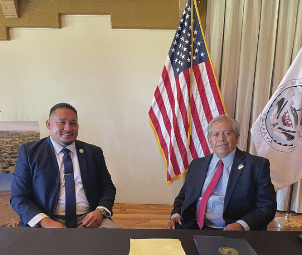 Two seated men in suits sitting at a table in front of a US flag and a tribal flag.