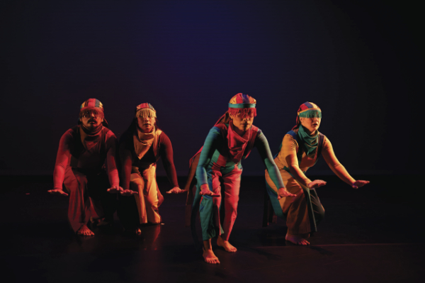 Four dancers performing on a stage with a black drop. The dancers are wearing red, turquoise, and yellow costumes with strong vertical stripes and head pieces.