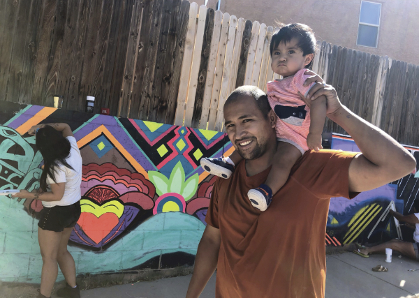 Man holding baby on his shoulders walking by a vibrant mural being painted by a woman with long hair
