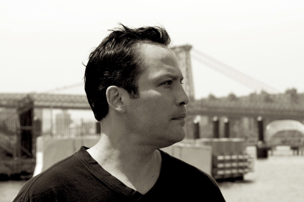 Sepia toned profile portrait of a man in front of a waterfront with bridge.