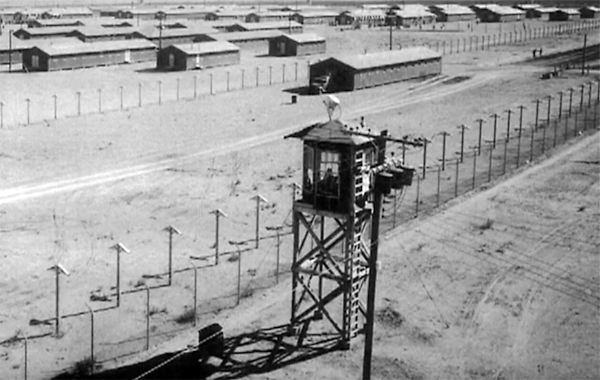 black and white vintage photograph of camp watchtower and perimeter fenching with bunks in the distance