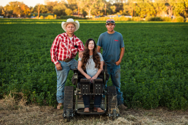 A woman posing in her wheelchair amidst the fields of her farm flanked by her husband and son