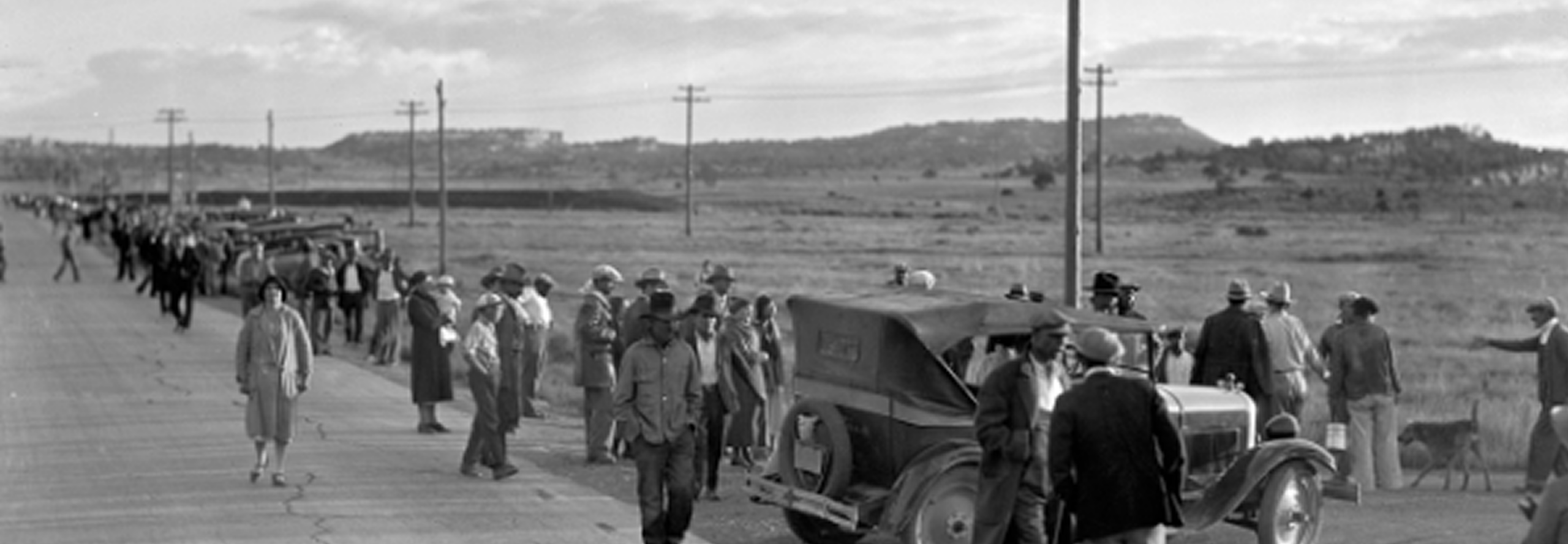 A large group of people, some standing by vintage cars and others walking in a line along a rural road, with open fields and telephone poles in the background.