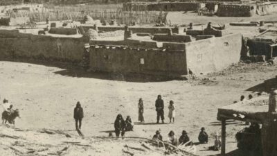 Historic photograph of an adobe village with several people standing or walking in an open central area, surrounded by flat-roofed buildings and fenced enclosures. [gen-ai]