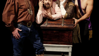 Six adults in theatrical costumes pose dramatically around a wooden chest onstage; expressions and attire suggest a comedic or whimsical performance. [gen-ai]