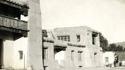 Black-and-white photo of adobe-style buildings with wooden beams, a balcony, and a few people standing near the structures; mountains are visible in the distance. [gen-ai]