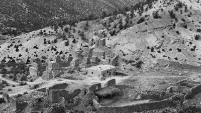 Black and white photo of ancient stone ruins situated on a hillside, surrounded by sparse trees and rocky terrain. [gen-ai]