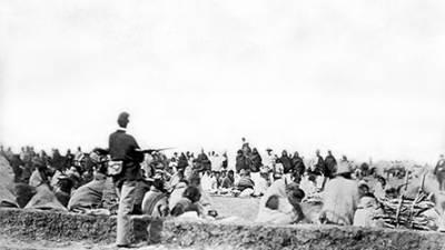 Black-and-white photo of a soldier with a rifle standing guard over a large group of seated prisoners or detainees in an outdoor camp setting. [gen-ai]