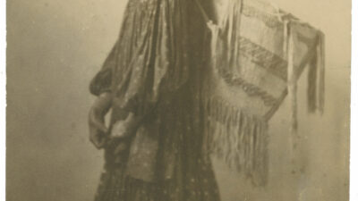 Historic sepia photograph of a young Indigenous woman in a patterned dress, standing in profile, carrying a woven basket or cradleboard on her back. [gen-ai]