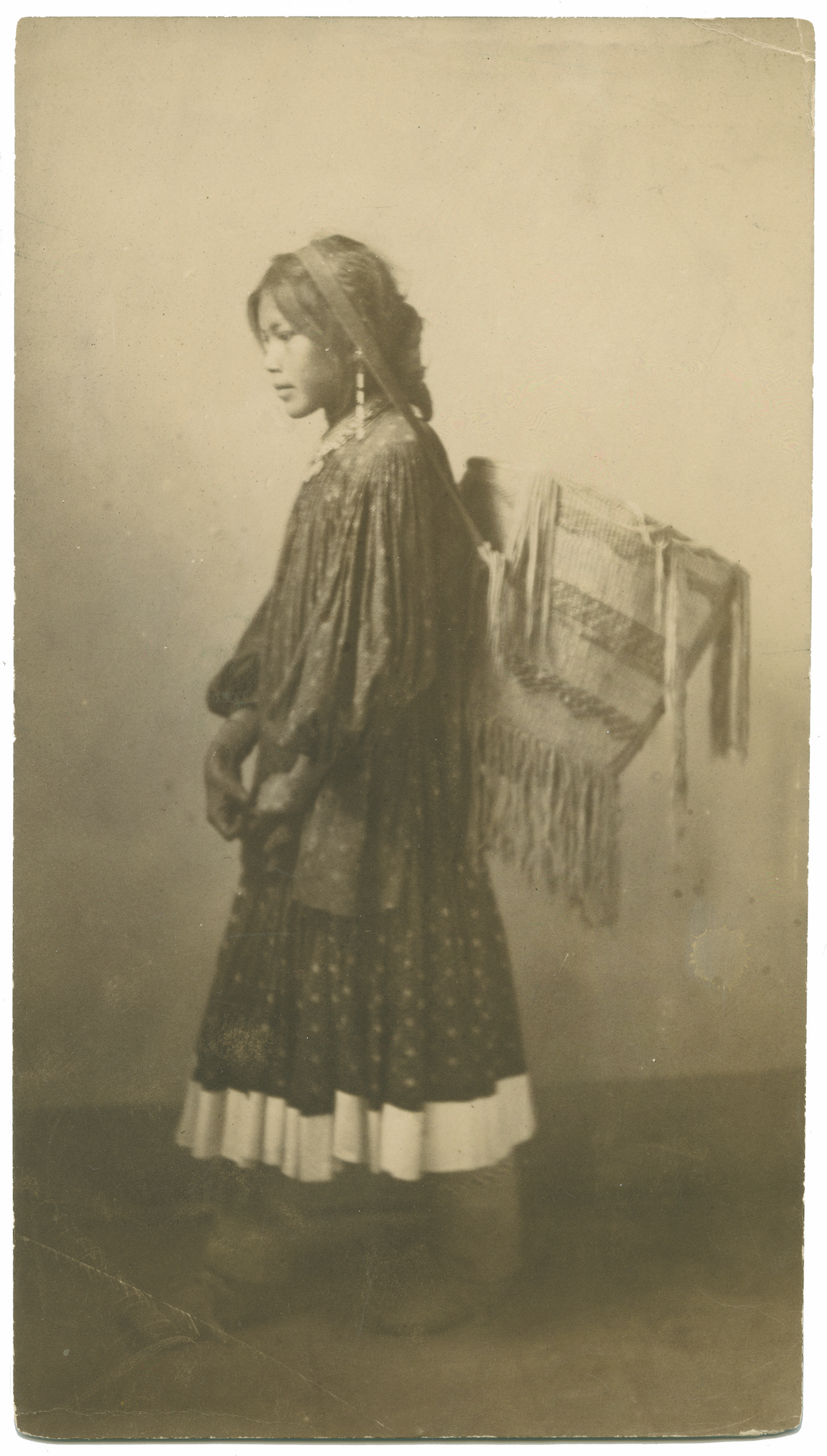 Historic sepia photograph of a young Indigenous woman in a patterned dress, standing in profile, carrying a woven basket or cradleboard on her back. [gen-ai]