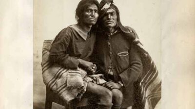 Two Navajo men sit side by side; one wears a dark shirt and skirt, the other a jacket and blanket, both looking toward the camera. Handwritten text above reads, Navajo Chiefs. [gen-ai]