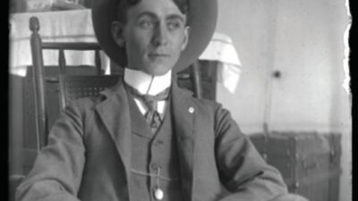 A young man wearing a suit, tie, and wide-brimmed hat sits in a wooden chair, with a table and other furniture visible in the background. [gen-ai]