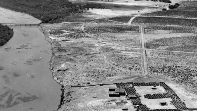 Aerial view of an arid landscape with a river on the left, a bridge in the distance, and a fenced compound containing buildings and vehicles on the right. [gen-ai]