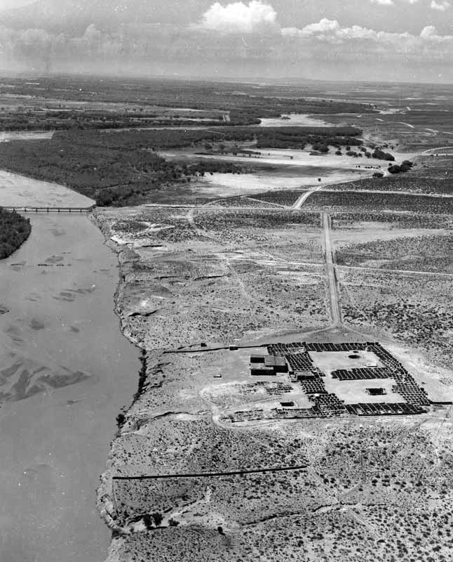 Aerial view of an arid landscape with a river on the left, a bridge in the distance, and a fenced compound containing buildings and vehicles on the right. [gen-ai]