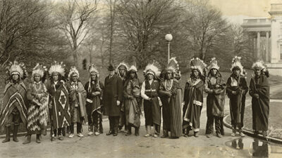 A group of Native American men in traditional attire and feather headdresses stand in a line outdoors with a man in a suit; trees and a large building are in the background. [gen-ai]