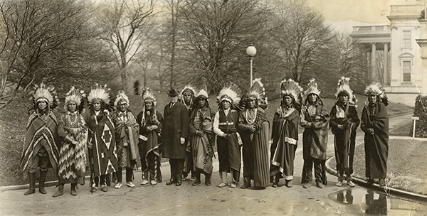 A group of Native American men in traditional attire and feather headdresses stand in a line outdoors with a man in a suit; trees and a large building are in the background. [gen-ai]