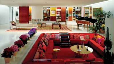 A sunken red seating area with patterned cushions is surrounded by potted flowers, a gold table, bookshelves, a grand piano, and modern decor in a spacious living room. [gen-ai]