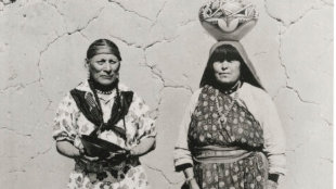 Two Indigenous women stand side by side against a cracked wall, one holding a bowl and the other with a woven basket balanced on her head. [gen-ai]