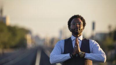 A man wearing glasses and a vest stands on railway tracks with hands pressed together in a prayer gesture. [gen-ai]