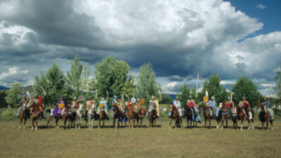 A group of people in colorful attire sit on horseback in a row on a grassy field with trees and dramatic clouds in the background. [gen-ai]