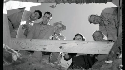 Six people stand above and look down into a pit, framed by wooden beams and a rope pulley system, under a clear sky. [gen-ai]