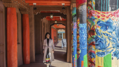 A woman in a long coat walks under a covered walkway with brightly painted pillars and adobe-style architecture. [gen-ai]