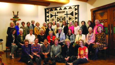 A group of women and a few men pose together indoors in front of a patterned wall hanging, sitting and standing in rows, smiling at the camera. [gen-ai]