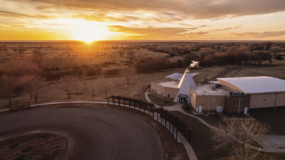 A building with a conical roof stands near an arena at sunset, surrounded by trees and open fields under a partly cloudy sky. [gen-ai]