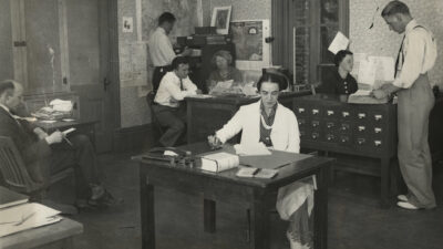 Black-and-white photo of six people working in an office with desks, papers, filing cabinets, and office equipment, likely from the early to mid-20th century. [gen-ai]