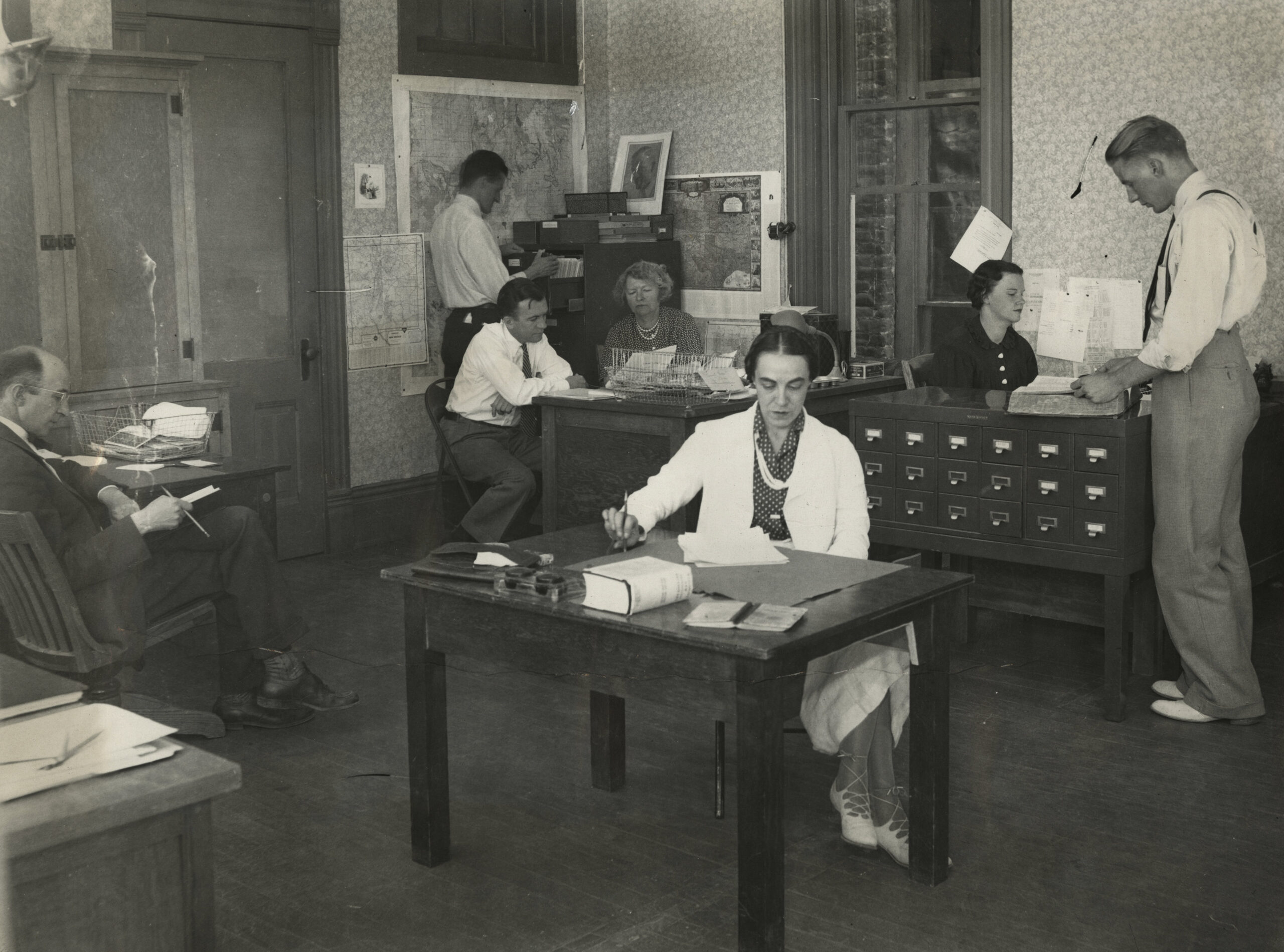Black-and-white photo of six people working in an office with desks, papers, filing cabinets, and office equipment, likely from the early to mid-20th century. [gen-ai]