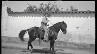 A person wearing a sombrero rides a dark horse on a street beside a white stucco wall with a tiled roof. [gen-ai]