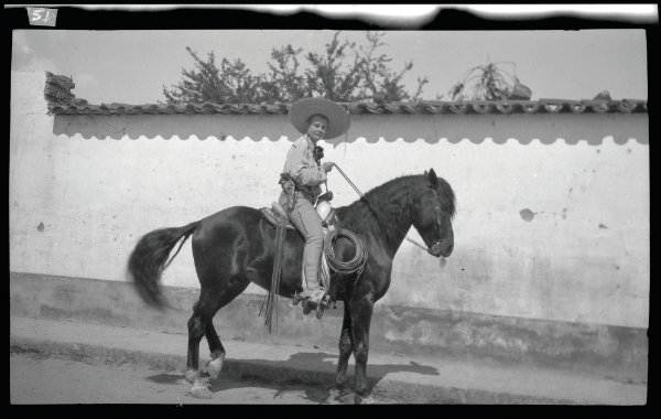 A person wearing a sombrero rides a dark horse on a street beside a white stucco wall with a tiled roof. [gen-ai]