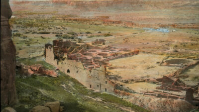 A view of ancient stone ruins in a desert landscape, partially surrounded by low vegetation and rocky terrain, with distant cliffs and a river in the background. [gen-ai]