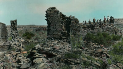 Several people stand atop and beside crumbling stone ruins in a rocky, overgrown landscape under a partly cloudy sky. [gen-ai]