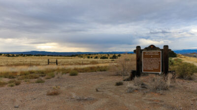 A brown historical marker sign stands beside a rural road, overlooking a wide, dry grassy plain under a cloudy sky. [gen-ai]