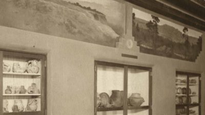 Sepia-toned photo of a museum room with shelves displaying pottery and artifacts, and landscape murals decorating the upper walls. [gen-ai]