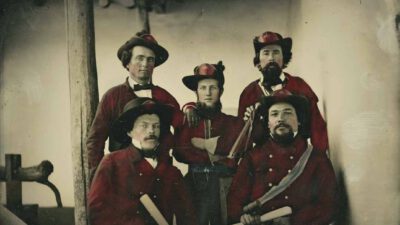 Five men in old-fashioned red uniforms pose indoors, four holding scrolls and one with a sword; the photo has a vintage, hand-colored appearance. [gen-ai]