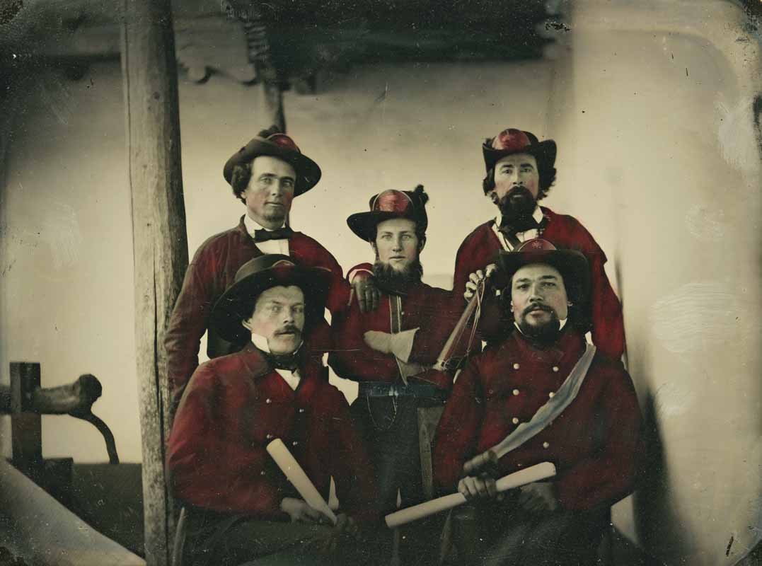 Five men in old-fashioned red uniforms pose indoors, four holding scrolls and one with a sword; the photo has a vintage, hand-colored appearance. [gen-ai]