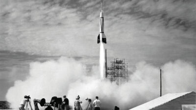 A rocket launches vertically as a group of people with cameras and equipment observe from the ground, surrounded by smoke and a partly cloudy sky. [gen-ai]