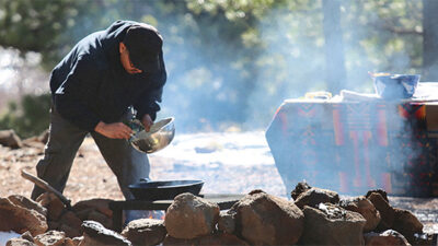 A person cooks over a campfire surrounded by rocks in a wooded outdoor setting, with a table covered by a patterned cloth in the background. [gen-ai]