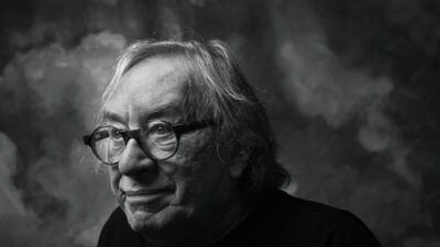 Black and white portrait of an older man with gray hair and glasses, wearing a dark shirt, looking slightly to the left against a cloudy textured background. [gen-ai]
