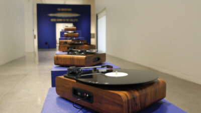 A row of wooden turntables with vinyl records are displayed on blue pedestals in a modern art gallery with a blue wall in the background. [gen-ai]