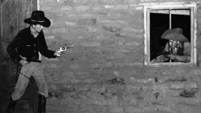 Two cowboys aiming guns at each other, one standing by a door and the other peering through a window in a stone building. [gen-ai]