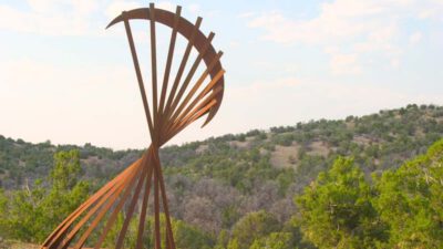 Large abstract metal sculpture with curved and radiating beams set outdoors on a hill, surrounded by green trees and a distant forested landscape. [gen-ai]