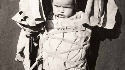A baby wrapped in a traditional Native American cradleboard, with fabric and leather coverings, against a plain background. [gen-ai]