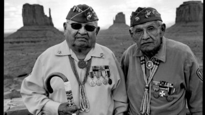 Two elderly men in military uniforms with medals stand in front of rock formations in a desert landscape. [gen-ai]