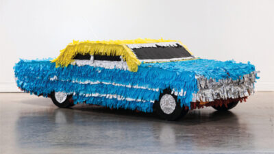 A life-sized car sculpture covered in blue, yellow, white, and silver fringe, resembling a piñata, displayed in a gallery setting. [gen-ai]
