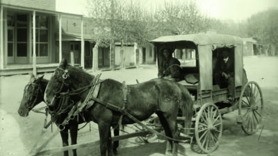 Two horses pull a covered wooden wagon carrying two people on a dirt road in front of old-fashioned buildings. [gen-ai]